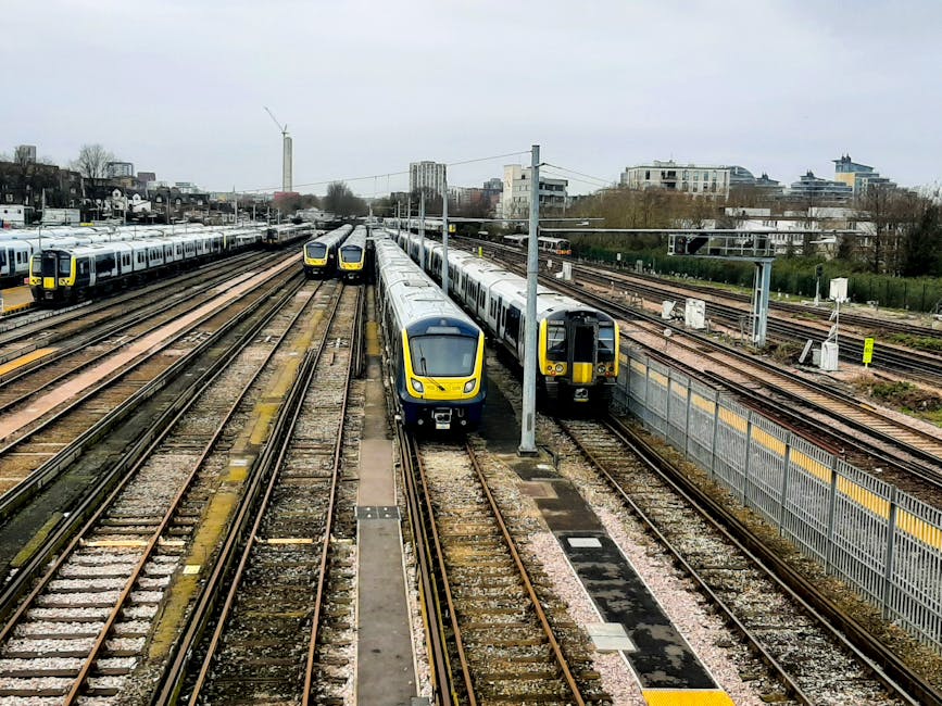 A view of multiple train tracks in a railway yard with two modern yellow and grey passenger trains positioned on adjacent tracks. The tracks are made of rust-colored rails mounted on gravel beds, with