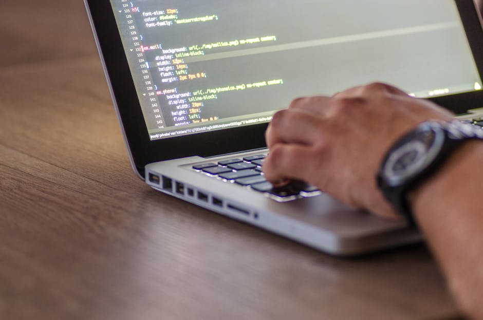 A close-up image of a person's hand typing on a silver laptop keyboard, displaying lines of color-coded code on the screen. The person's wrist is adorned with a black watch. The laptop is placed on a 